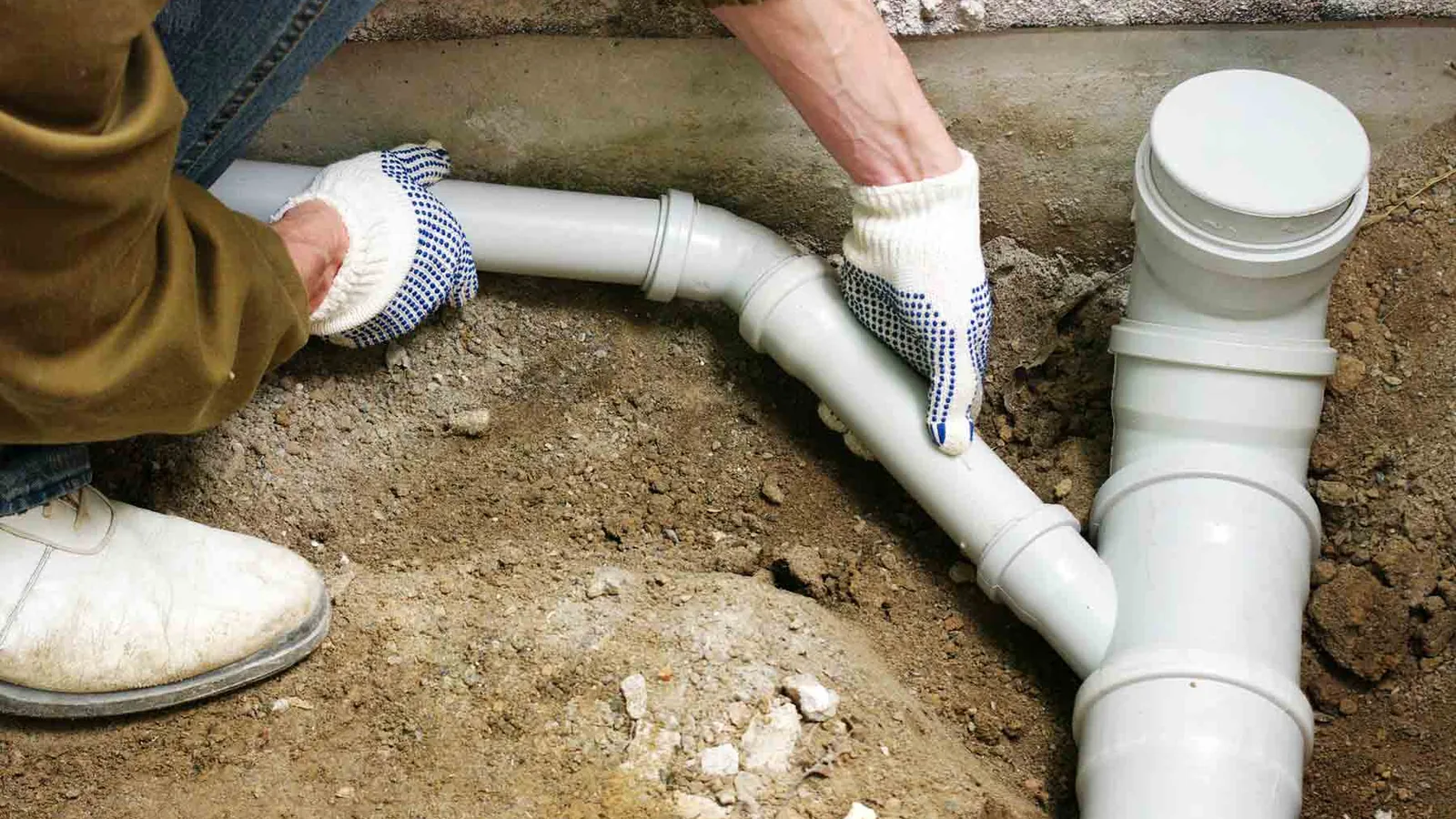 Gloved hands fitting new PVC drain pipe and a cleanout at a house foundation